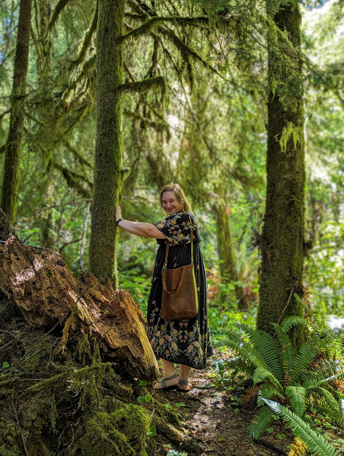 A full-length portrait of Stephy, a blonde white woman standing in a lush, sun-dappled temperate rainforest. She is leaning slightly, standing with her right hand extended to reach a moss-covered tree trunk. She is wears a long, flowing, dark-colored dress with a folksy botanical print and carries a large, cognac-brown leather shoulder bag. The forest is thick with vibrant green ferns, mosses, and high, tall trees whose trunks are heavily draped in moss and foliage, giving the scene an ethereal, 'forest witch' aesthetic. A shaft of bright sunlight breaks through the canopy onto Stephy and the forest floor.